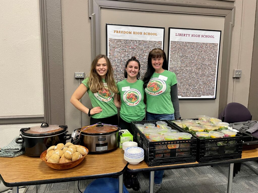Dr. Grega and volunteers standing behind a table smiling. Their are fresh and healthy mels prepared in crockpots to be served to community members.